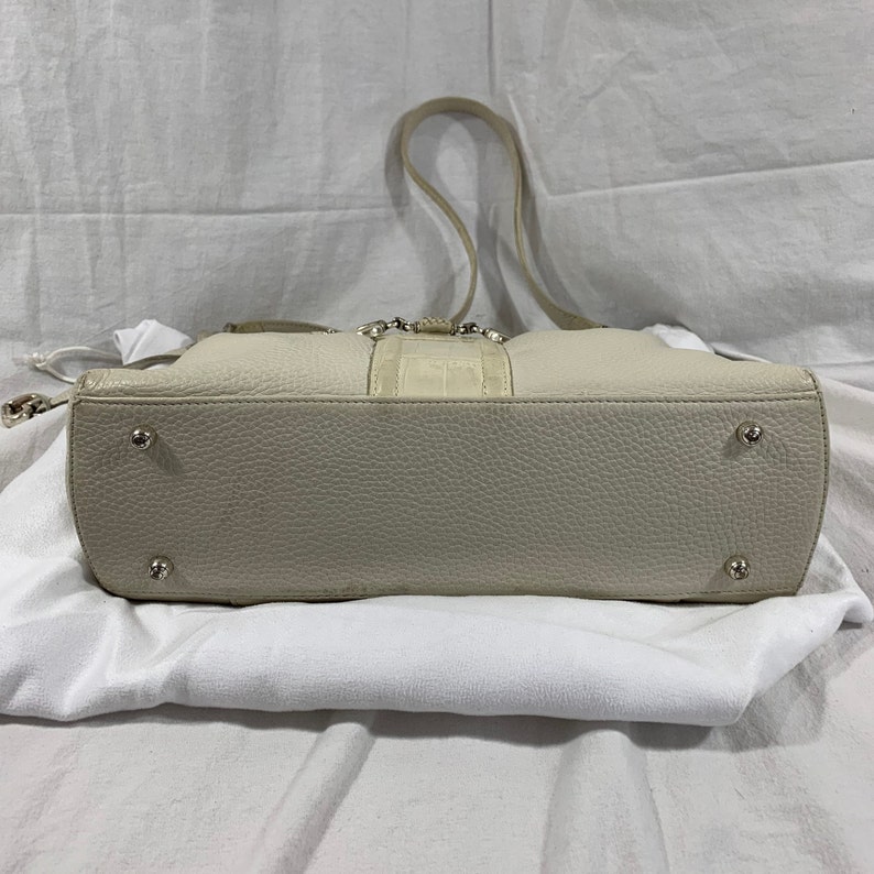 May include: A cream-colored leather tote bag with a crocodile embossed detail on the front. The bag has a top zipper closure and a single strap. The bag is sitting on a white surface.
