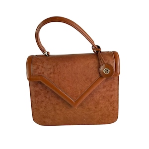 Op de afbeelding: Een vintage bruine handtas met een handvat aan de bovenkant en een getextureerde buitenkant. De tas heeft een geometrische klepsluiting en een klein, rond embleem aan de voorkant. De handgreep is gebogen.