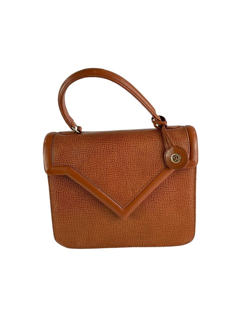 Op de afbeelding: Een vintage bruine handtas met een handvat aan de bovenkant en een getextureerde buitenkant. De tas heeft een geometrische klepsluiting en een klein, rond embleem aan de voorkant. De handgreep is gebogen.