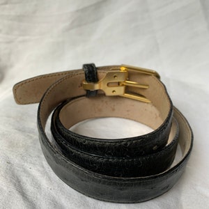 Op de afbeelding: Een zwarte leren riem met een gouden gesp. De riem is opgerold en de gesp is zichtbaar.
