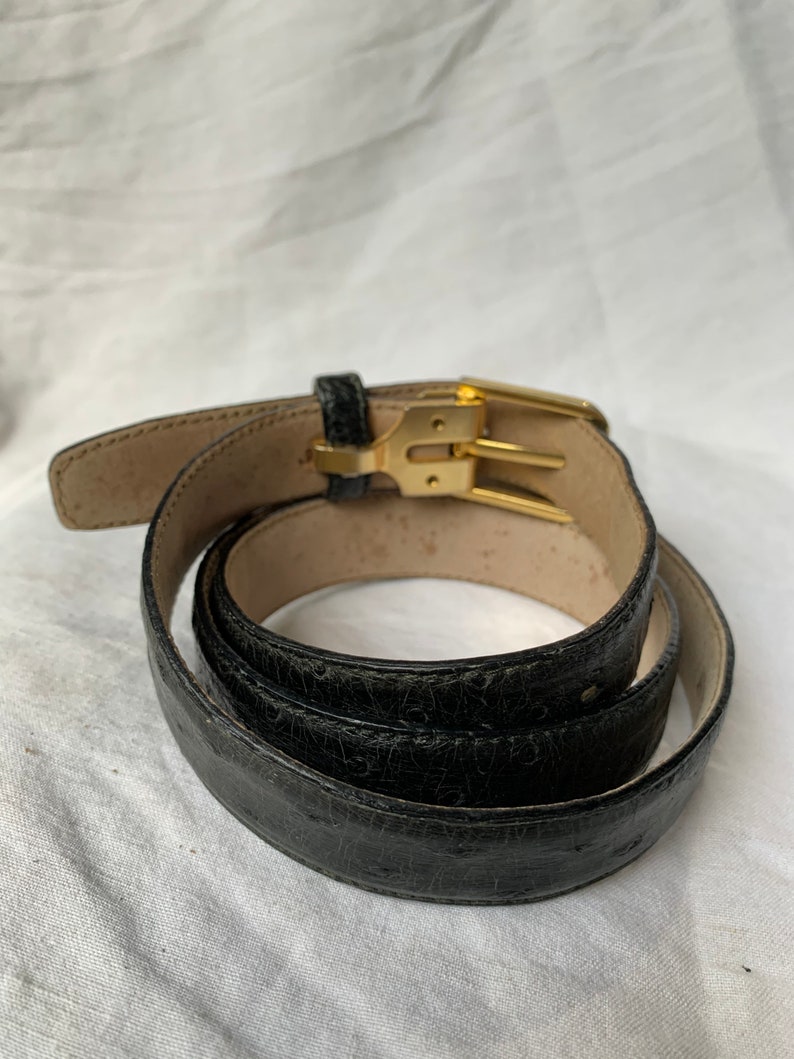 Op de afbeelding: Een zwarte leren riem met een gouden gesp. De riem is opgerold en de gesp is zichtbaar.