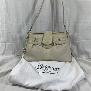 May include: A white leather Brighton handbag with a crocodile embossed detail and silver hardware. The bag has a top flap closure and a single strap. The bag is sitting on a white dust bag with the Brighton logo.