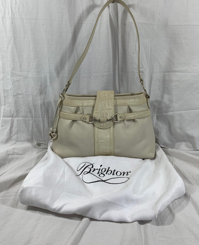 May include: A white leather Brighton handbag with a crocodile embossed detail and silver hardware. The bag has a top flap closure and a single strap. The bag is sitting on a white dust bag with the Brighton logo.