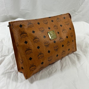 May include: A brown leather cosmetic bag with a black and brown monogram pattern. The bag has a gold metal logo with the letters MCM.