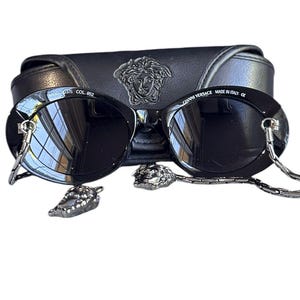 May include: Black round sunglasses with a silver chain attached to the frames. The sunglasses are in a black case with the Versace logo.