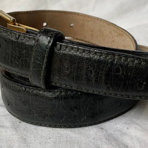 Op de afbeelding: Een zwarte leren riem met een gouden gesp. De riem heeft een getextureerd, gegraveerd patroon.