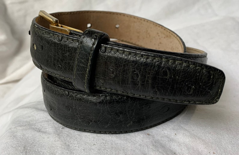 Op de afbeelding: Een zwarte leren riem met een gouden gesp. De riem heeft een getextureerd, gegraveerd patroon.
