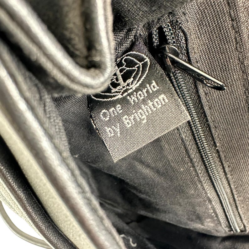 Op de afbeelding: Close-up van de binnenkant van een zwarte handtas met een rits en een label met de tekst "One World by Brighton". Het label heeft een wereldbolontwerp. Het materiaal van de tas lijkt leer of een leerachtig materiaal te zijn.