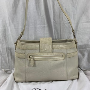 May include: A white leather Brighton shoulder bag with a crocodile embossed flap and a zippered pocket on the front. The bag is sitting on a white fabric surface with the Brighton logo.