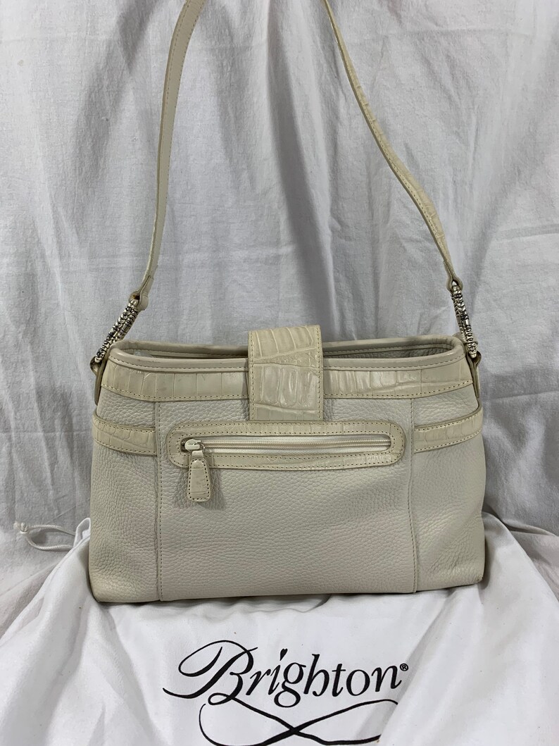 May include: A white leather Brighton shoulder bag with a crocodile embossed flap and a zippered pocket on the front. The bag is sitting on a white fabric surface with the Brighton logo.