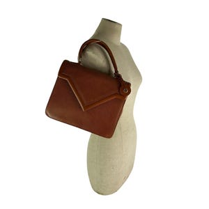 Op de afbeelding: Een bruine leren handtas met een gestructureerd ontwerp. De tas heeft een handvat aan de bovenkant en een klepsluiting. Het leer heeft een getextureerde uitstraling. De tas wordt op een mannequin getoond.