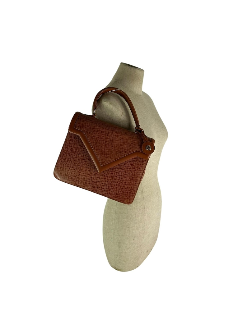 Op de afbeelding: Een bruine leren handtas met een gestructureerd ontwerp. De tas heeft een handvat aan de bovenkant en een klepsluiting. Het leer heeft een getextureerde uitstraling. De tas wordt op een mannequin getoond.