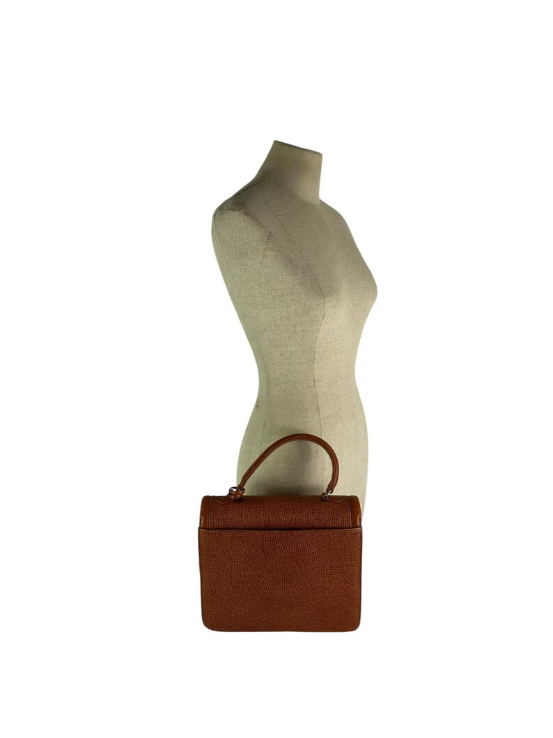 Op de afbeelding: Een bruine leren handtas met een handvat aan de bovenkant en een rechthoekige vorm. De tas heeft een textuuroppervlak en een klepsluiting. De tas wordt op een mannequin getoond.