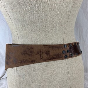 May include: A brown leather belt with metal studs. The belt is worn on a dress form.