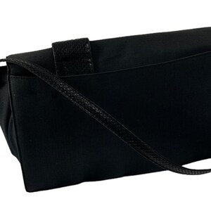 May include: Black messenger bag with a textured flap closure and a woven shoulder strap. The bag has a rectangular shape with a flap that secures with a textured leather strap. The shoulder strap is thin and woven.