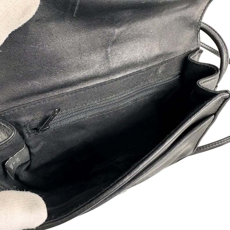 Op de afbeelding: Open zicht op een zwarte leren crossbodytas. De tas heeft een klepsluiting en een lange, dunne riem. De binnenkant heeft een vak met ritssluiting en een ruim hoofdvak. De tas is ontworpen met een minimalistische esthetiek.