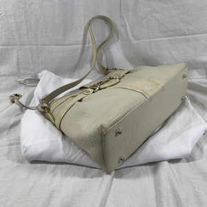 May include: A white leather handbag with a silver buckle and a long strap. The bag is sitting on a white surface.