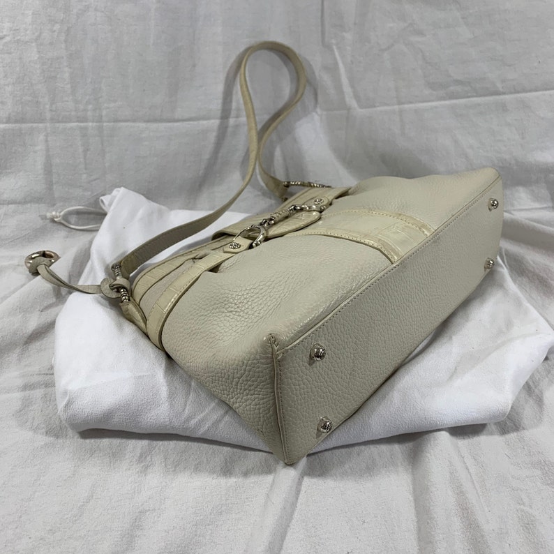 May include: A white leather handbag with a silver buckle and a long strap. The bag is sitting on a white surface.