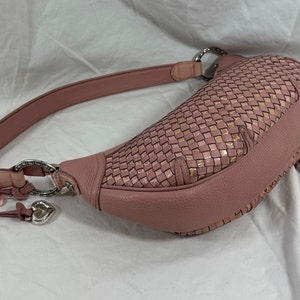 May include: A dusty rose-colored hobo bag with a woven top section and a leather strap. The bag features silver-tone hardware, a heart-shaped charm, and a small tassel detail. The bag is designed with a curved shape.