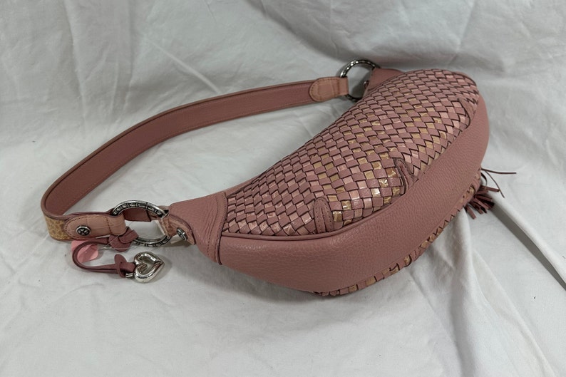 May include: A dusty rose-colored hobo bag with a woven top section and a leather strap. The bag features silver-tone hardware, a heart-shaped charm, and a small tassel detail. The bag is designed with a curved shape.