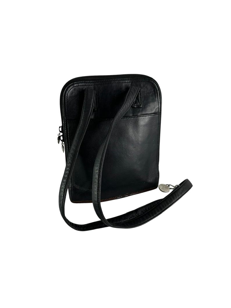 May include: Black leather backpack with adjustable straps and a top zipper closure. The bag has a rectangular shape with rounded corners and a small silver-toned charm attached to the strap.