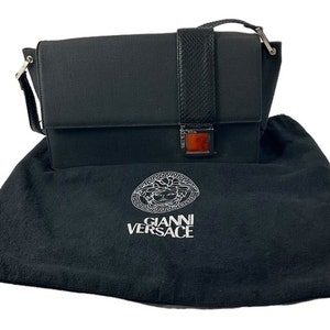 Może przedstawiać: Czarna prostokątna torebka z klapką i paskiem o fakturze skóry węża. Torebka ma srebrne zapięcie z prostokątnym bursztynowym akcentem. Leży na czarnym worku przeciwkurzowym z logo "GIANNI VERSACE".