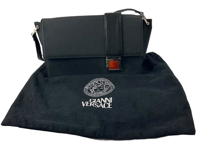May include: Black rectangular handbag with a flap closure and a snakeskin-textured strap. The bag has a silver-tone clasp with a rectangular amber-colored accent. It rests on a black dust bag with the "GIANNI VERSACE" logo.