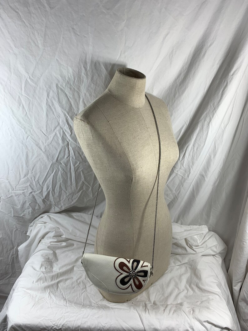 May include: A white leather half-moon shaped crossbody bag with a brown and white floral design and a silver chain strap.