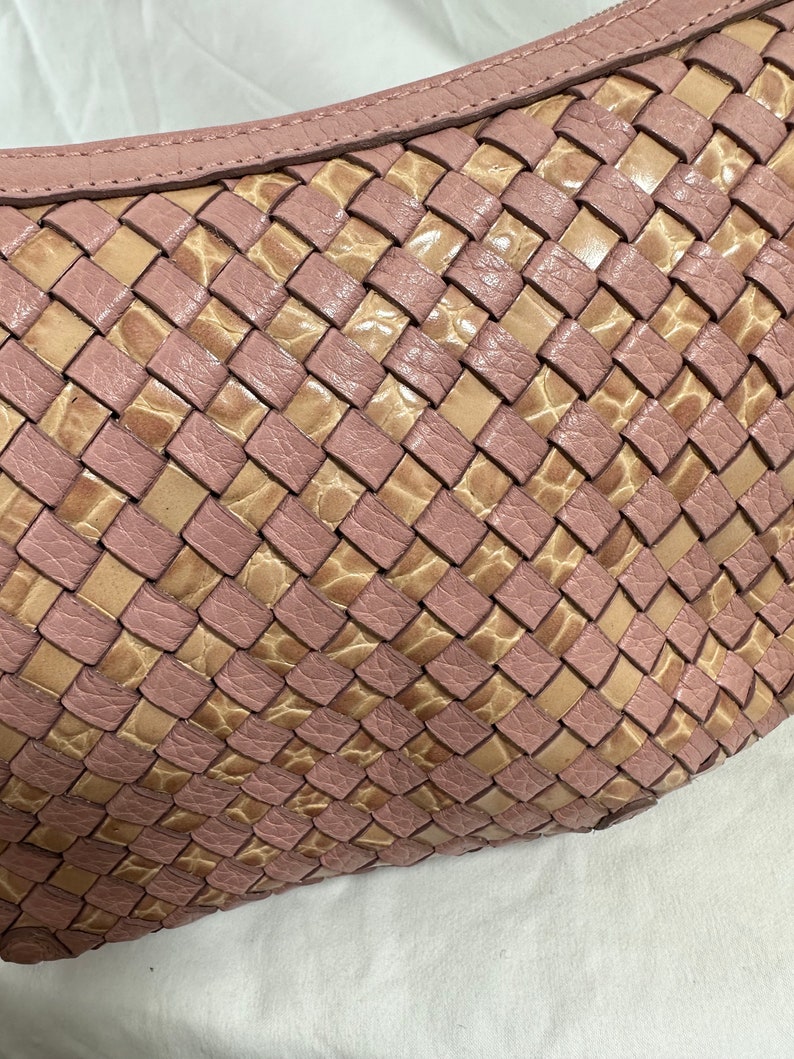 May include: Close-up of a woven leather handbag in a soft pink and tan color scheme. The bag features a basketweave design with a curved top and a subtle texture. The leather appears to be high-quality.