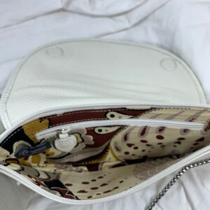 May include: White leather purse with a chain strap and a patterned fabric lining. The purse has a zipper closure and a small metal charm.