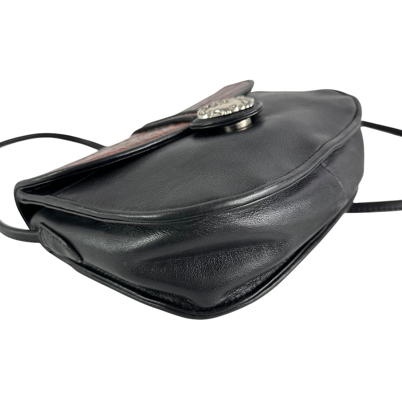 Op de afbeelding: Zwarte leren crossbodytas met een decoratieve zilverkleurige sluiting. De tas heeft een gebogen ontwerp en een bruine en patroonklep. De riem is dun en zwart, geschikt om over de schouder of crossbody te dragen.