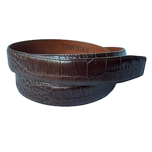 May include: A dark brown belt with a glossy crocodile-embossed texture. The interior of the belt is a smooth tan leather and features the text TERRY STACK printed in black. The belt is coiled into a circular shape, showing the outer pattern.
