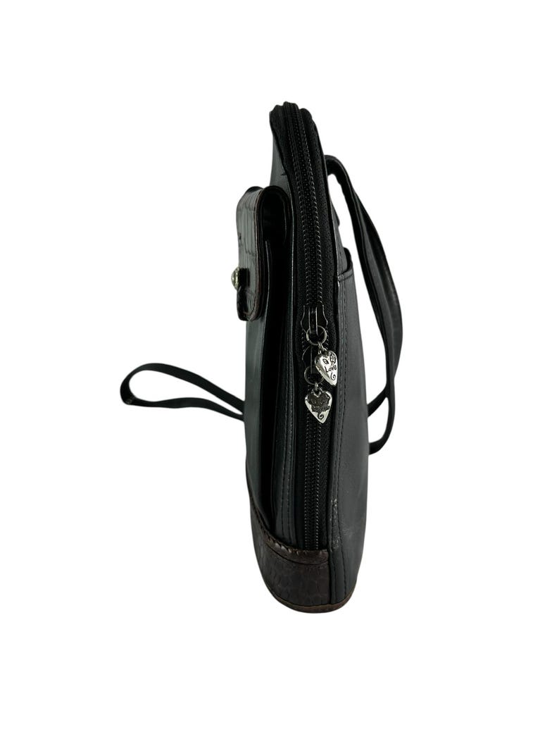 May include: Black leather crossbody bag with a brown leather bottom and a small front pocket. The bag has a zipper closure with heart-shaped charms and a long strap for wearing across the body.