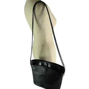 May include: Black leather crossbody bag with a textured, curved top and a braided strap. The bag is displayed on a mannequin and has a silver-toned clasp. The bag is a classic style.