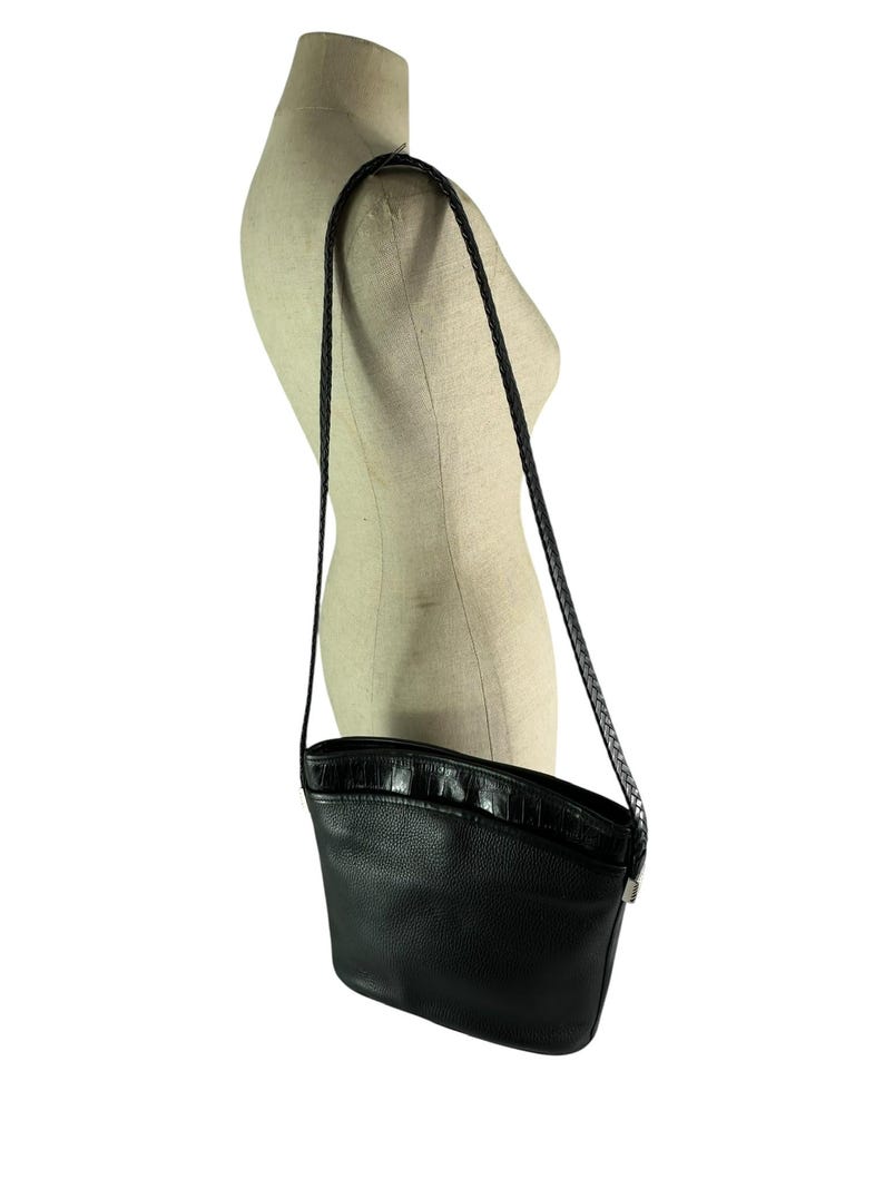 May include: Black leather crossbody bag with a textured, curved top and a braided strap. The bag is displayed on a mannequin and has a silver-toned clasp. The bag is a classic style.