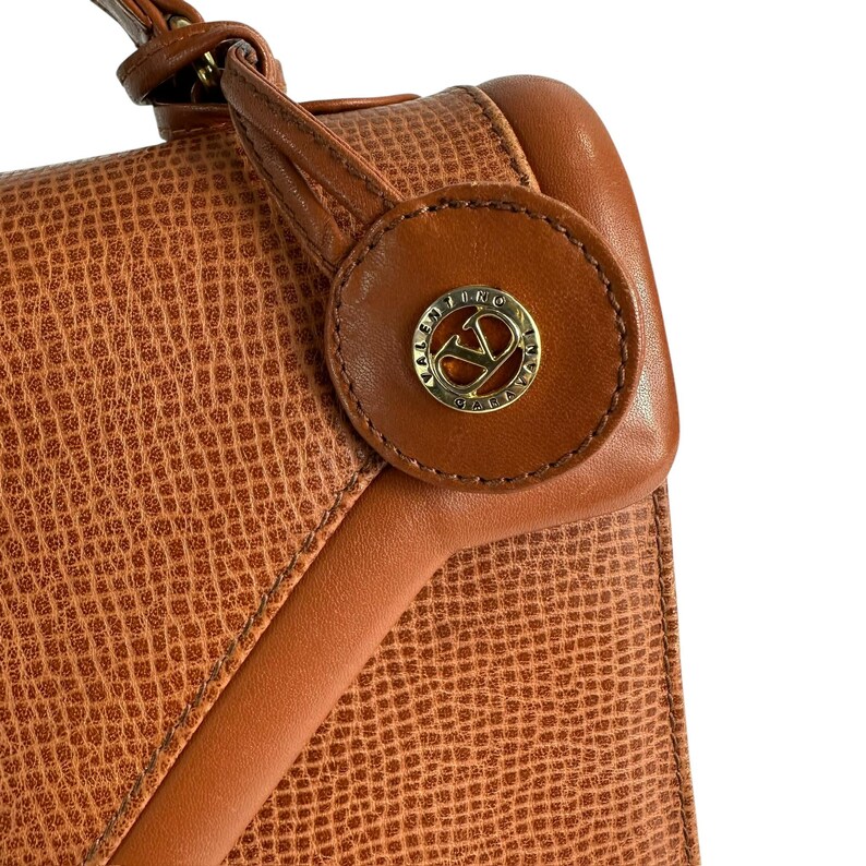 Op de afbeelding: Close-up van een bruine leren handtas met een textuur van slangenleer. De tas heeft een ronde, bruine lederen accent met een gouden logo. De tas heeft een handvat aan de bovenkant en een klepsluiting.