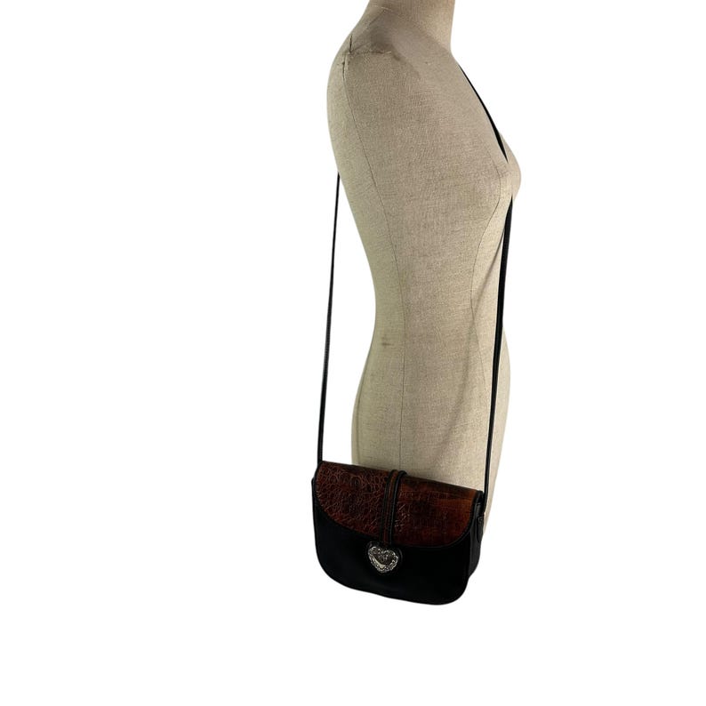 Op de afbeelding: Zwarte crossbodytas met een bruine, getextureerde flap en een zilverkleurige hartvormige sluiting. De tas heeft een lange, dunne zwarte riem. De tas wordt op een mannequin gepresenteerd.