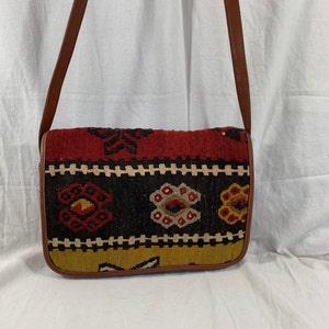 May include: A brown leather crossbody bag with a colorful woven tapestry design. The tapestry features geometric patterns in shades of red, yellow, black, and white.