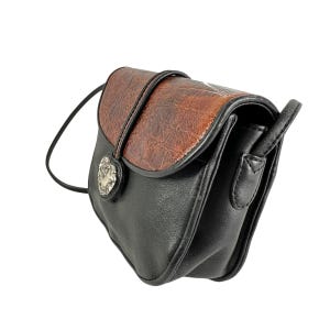 Op de afbeelding: Een zwarte en bruine crossbodytas met een getextureerde flap en een zilverkleurige hartvormige sluiting. De tas heeft een lange, dunne riem en is gemaakt van imitatieleer. De afmetingen van de tas zijn ongeveer 20 cm breed, 18 cm hoog en 8 cm diep.