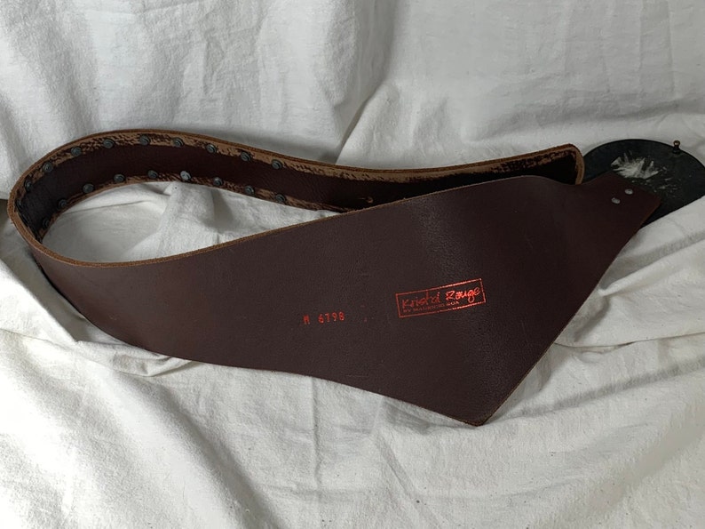 May include: A brown leather belt with a metal buckle and rivets. The belt is stamped with the text "Knotted Rouge" and the number "M 6795".