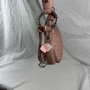 May include: A dusty rose-colored handbag with a woven design. The bag features a silver-toned ring handle attachment and a decorative heart charm. The strap is a combination of pink and textured tan leather.