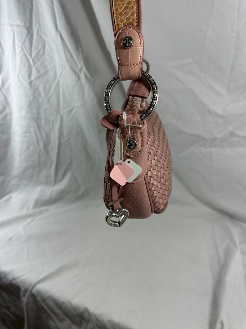 May include: A dusty rose-colored handbag with a woven design. The bag features a silver-toned ring handle attachment and a decorative heart charm. The strap is a combination of pink and textured tan leather.
