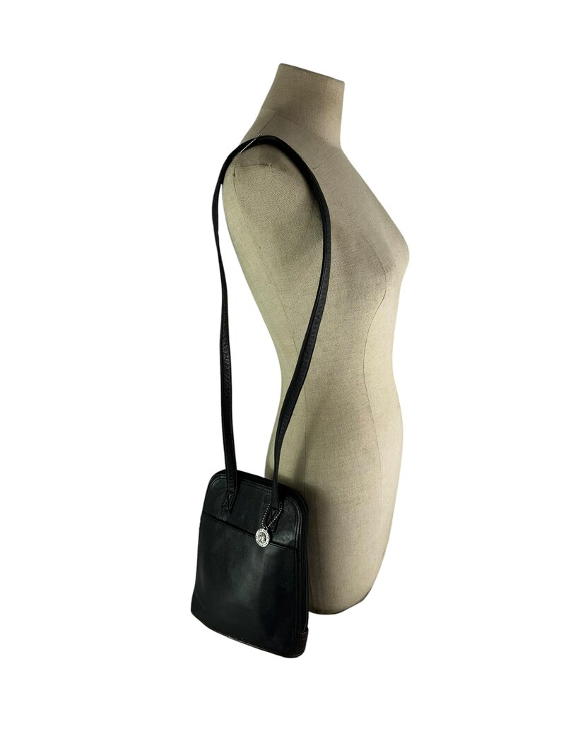 May include: Black leather shoulder bag with a long strap. The bag has a rectangular shape with rounded corners and a small silver charm. The strap is thin and appears to be adjustable. The bag is displayed on a mannequin.