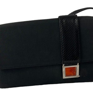 May include: Black messenger bag with a flap closure. The bag features a textured black strap and a decorative rectangular clasp with an orange-red center. The bag has a shoulder strap and a silver-colored metal buckle.