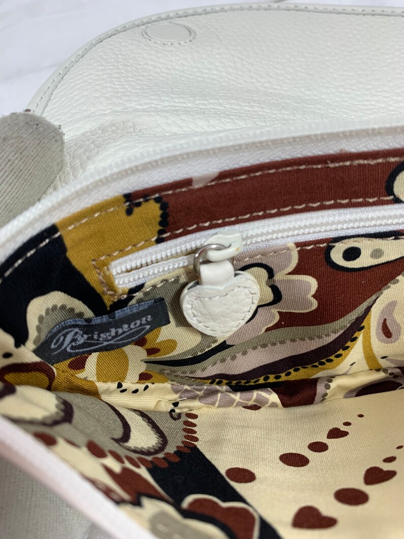 May include: The inside of a white leather purse with a paisley patterned lining. The purse has a zipper closure and a small leather tag that says "Brighton".
