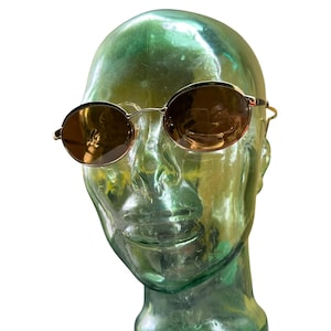 May include: Oval-shaped sunglasses featuring thin gold-tone metal frames and brown-tinted lenses. The eyewear is displayed on a translucent green mannequin head, highlighting the slim bridge and slender temples of the frame design.