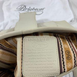May include: A white leather handbag with a brown and white striped lining. The tag inside the bag reads "The Brighton Collection handbags are individually crafted in China from quality leathers. Any markings on the surface are part of the natural beauty, texture and character of the leather, which make each bag unique."