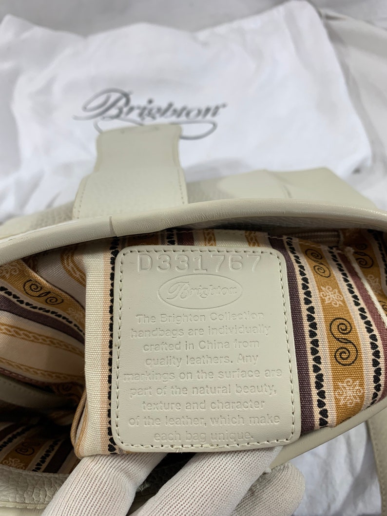 May include: A white leather handbag with a brown and white striped lining. The tag inside the bag reads "The Brighton Collection handbags are individually crafted in China from quality leathers. Any markings on the surface are part of the natural beauty, texture and character of the leather, which make each bag unique."