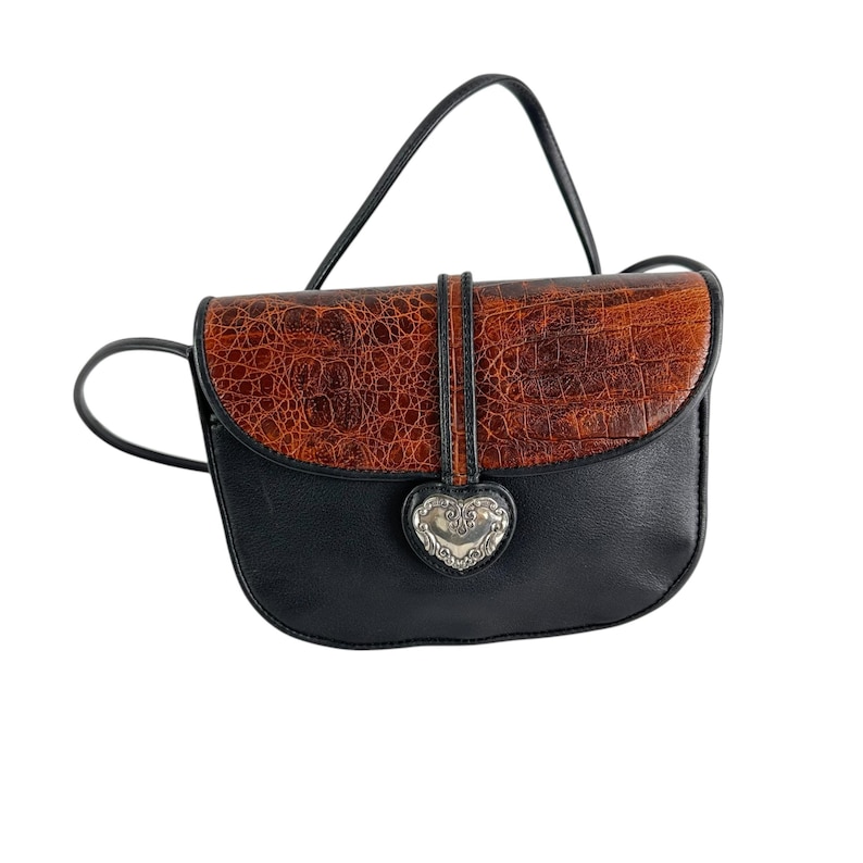 Op de afbeelding: Zwarte en bruine crossbodytas met een klep met textuur van imitatiekrokodil. De tas heeft een zilverkleurige hartvormige sluiting en een zwarte riem. De tas is ontworpen met een gebogen vorm en een enkele riem.