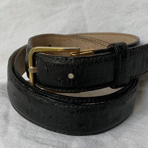Op de afbeelding: Een zwarte leren riem met een gouden gesp. De riem is gemaakt van struisvogelhuid en heeft een textuur.
